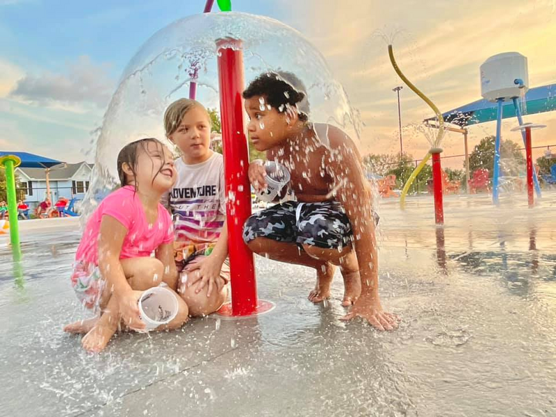 3 children at an aquatic playground park sit under a water feature Fun-Brella as the water shoots out of a column and creates a dome around them. water is splashing in the background of the spray park by splash pad equipment manufacturer Waterplay